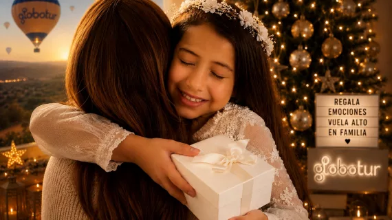 Niña vestida de Primera Comunión abrazando emocionada a un adulto mientras sostiene una caja de regalo blanca. Al fondo se ve un ambiente cálido y familiar con luces decorativas, un árbol iluminado y un globo aerostático de Globotur al atardecer. Un cartel luminoso muestra el mensaje “Regala emociones, vuela alto en familia